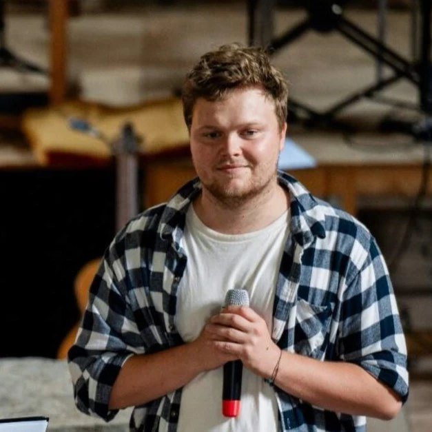 A man holding a microphone in his hand, wearing a white t-shirt and a blue and black checkered shirt.